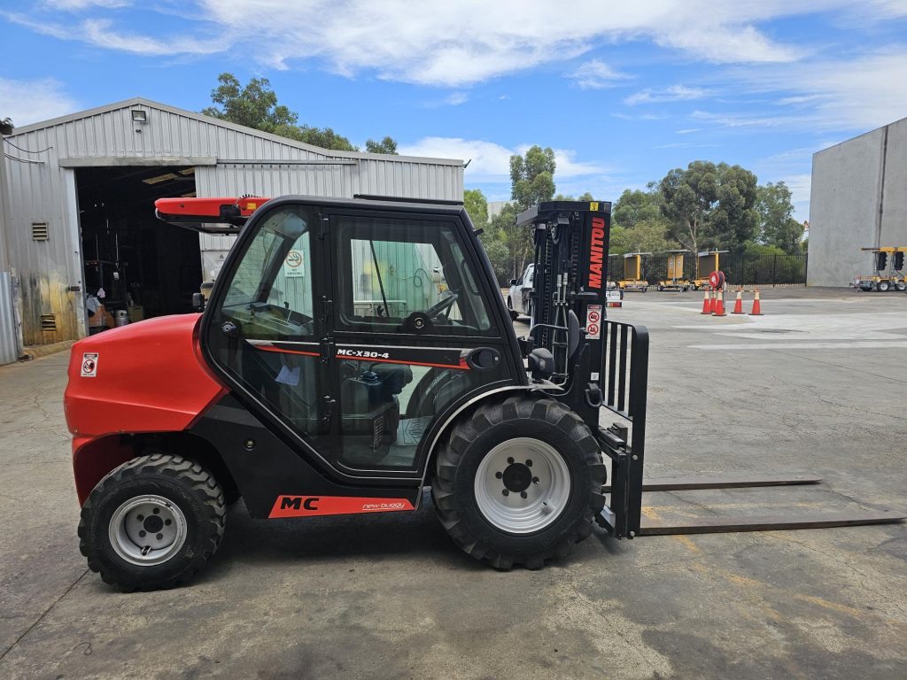 Red and black MC telehandler forklift parked in an industrial yard with warehouses and trees in the background.