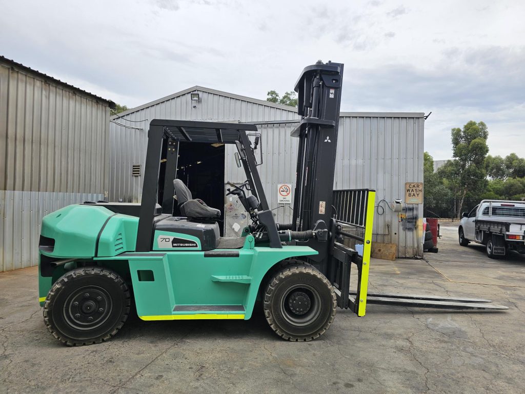 Turquoise Mitsubishi forklift with yellow forks parked outside industrial warehouse building.